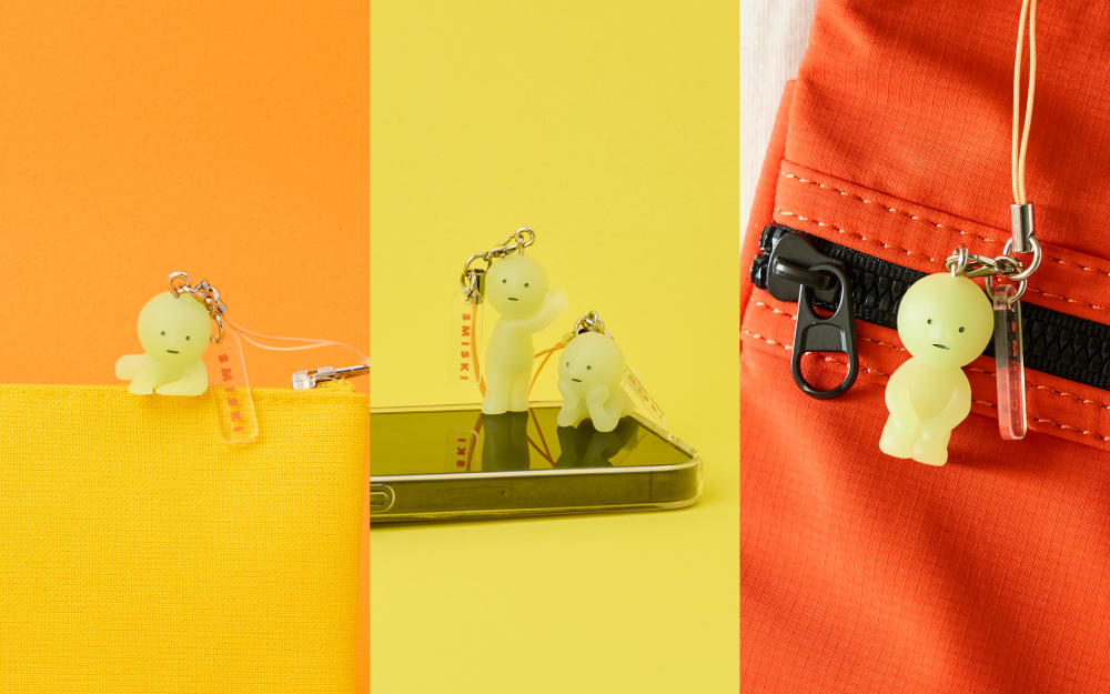 Split images shows new SMISKI accessory product. Left side image: set on a orange background, SMISKI Climbing is atop a yellow bag. Center: set against a bright yellow background, SMISKI Listening, and SMISKI Daydreaming variants stand and lay atop a cell phone. Right: image set against white background shows a red zippered bag with the SMISKI Kneeling variant attached.