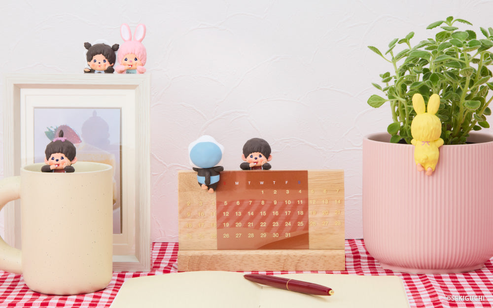 Monchhichi HIPPERS set on a checkered tablecloth with a mug, plant, and decorative items. From left to right, the girl figure is set in a cream colored mug, behind are two figures, the baby girl in hood, beside the pink bunny are set atop a framed image. In the center, two HIPPERS, the baby boy in hood facing away, and boy figure are set atop a wooden calendar. Far right, the yellow bunny figure is set atop a pink potted plant, facing away.