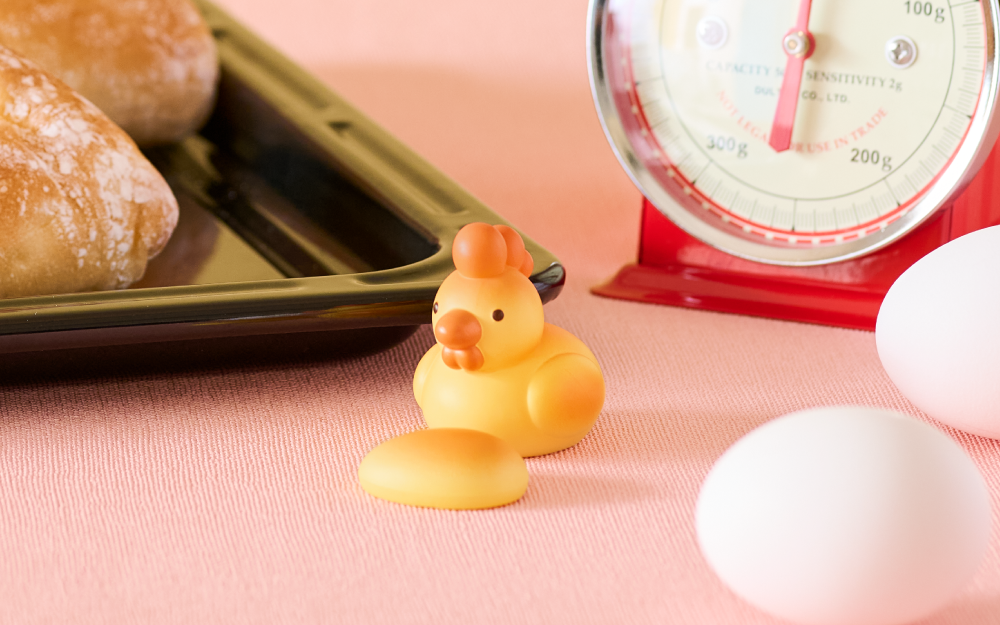 Yellow rubber duck with an orange hat on a pink surface with eggs and a scale in the background.